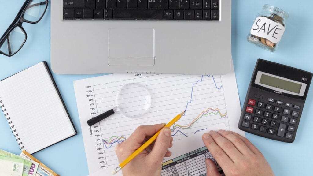 analista de finanças escrevendo em um papel com gráfico, sobre uma mesa com lupa, calculadora e notebook