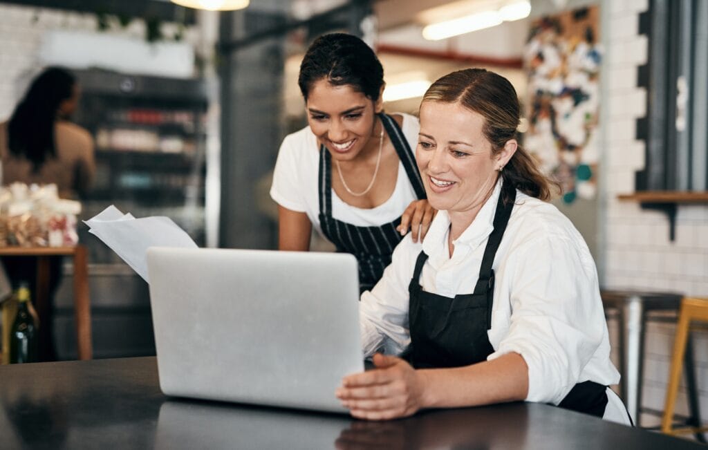 empreendedoras sentadas diante de um notebook com papeis sorrindo