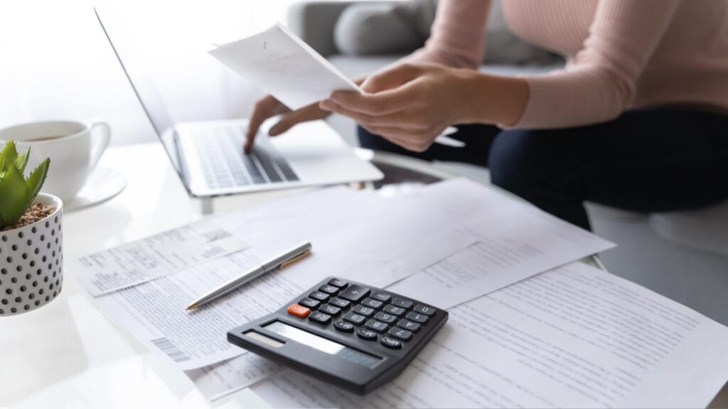empresária digitando no notebook e segurando um papel na outra mão. Na mesa há papeis e calculadora