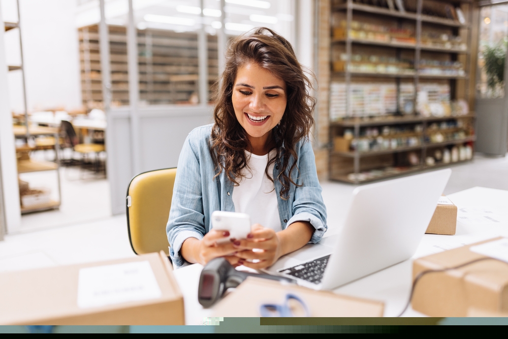 dona de loja mexendo no celular em frente ao notebook numa mesa